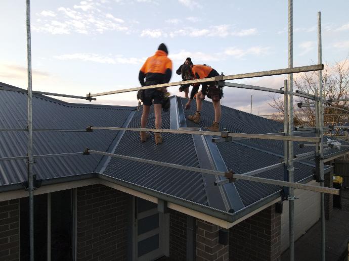 2 workers in orange hi-vis clothing installing a colorbond metal roof with safety scaffolding
